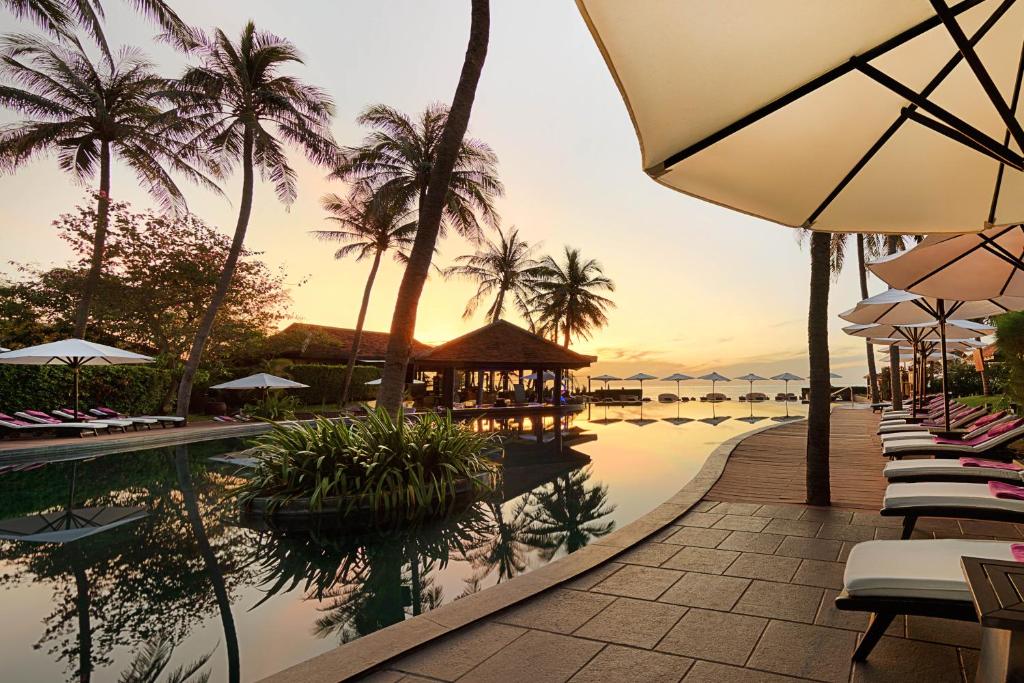 Tropu kūrorta baseins ar palmām un saulrietu fona, kas rada relaksējošu atmosfēru. Sauļošanās krēsli un saules vairogi pie baseina aicina uz atpūtu.