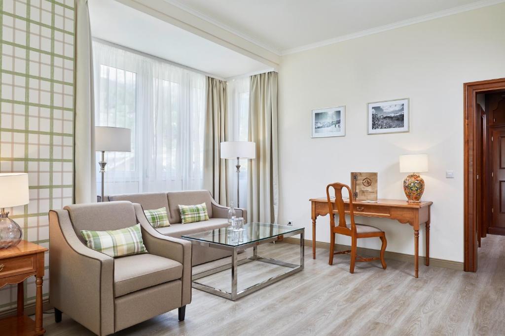 Mūsdienīgs viesistabas interjers ar gaišām krāsām, elegantu dīvānu un koka rakstāmgaldu. Logi ar vieglām aizkariem nodrošina dabīgu apgaismojumu un rada mājīgu atmosfēru.