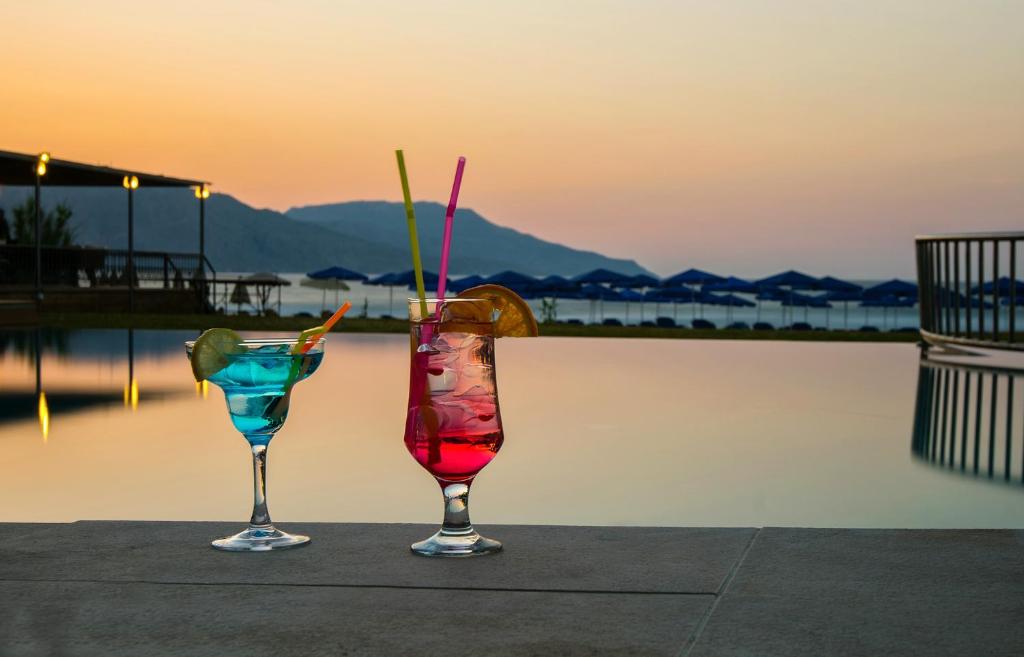 Divi krāsaini kokteiļi, viens zilā un otrs sarkanā krāsā, novietoti uz galda pie baseina ar skaistu saulrietu fona.