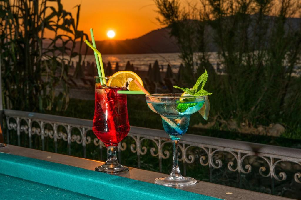 Divi krāsaini kokteiļi ar citrona un piparmētru dekorējumu, fona ainavā atspoguļojoties saulrietam pie jūras.