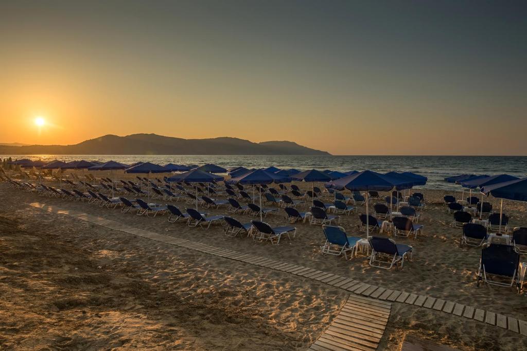Saule krīt pār pludmali, kur ir novietotas saules krēsli ar zilu jumtiņu. Skats uz jūru un kalniem veido mierīgu un relaksējošu atmosfēru.