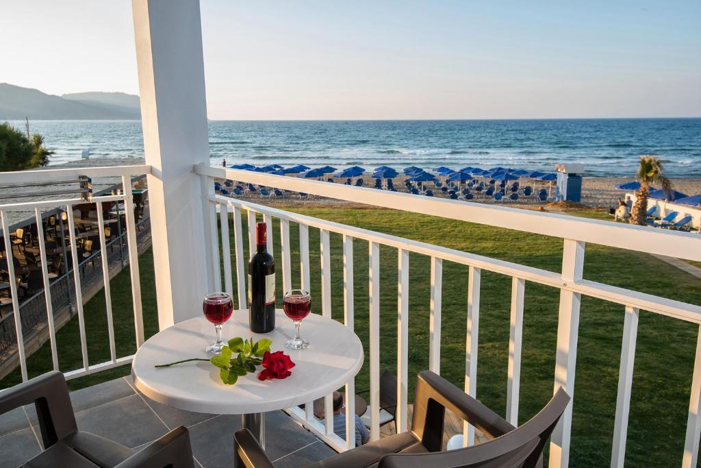 Romantiska skatu uz jūru no balkona, kur uz galda ir sarkanvīna pudele un divas glāzes ar sarkanvīnu, kā arī zieds. Fona ir pludmale ar saulessargiem un zāle.