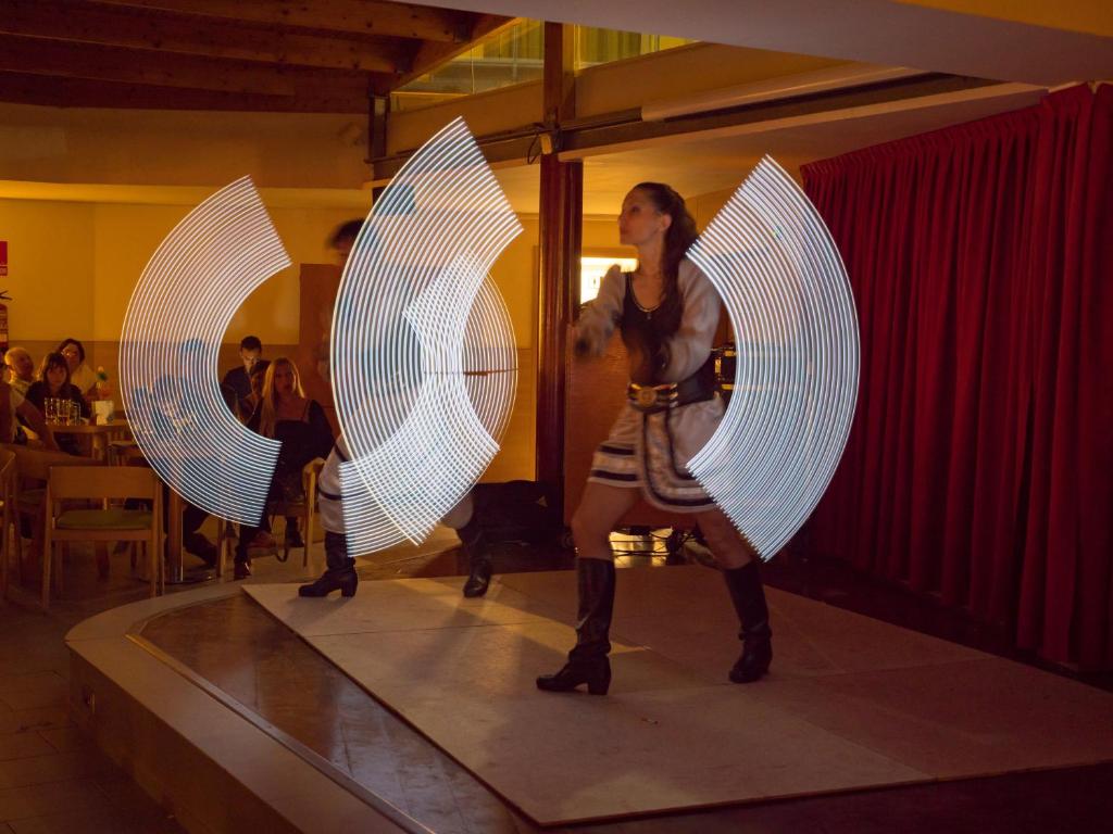 Gaismas priekšnesums ar dejotāju, kas izpilda kustības uz skatuves, radot spirālveida gaismas efektus. Fons ietver skatītājus, kas vēro izrādi.