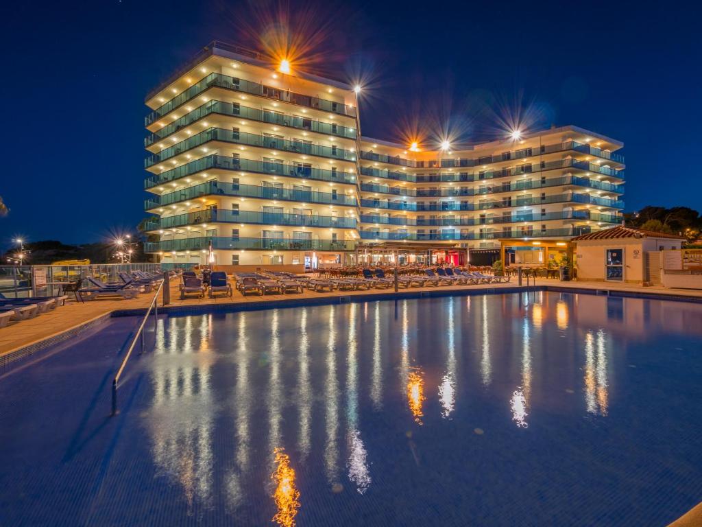 Moderns viesnīcas komplekss ar apgaismotām terasēm un baseinu, redzams tumšā vakarā. Viesnīcas ēka atspoguļojas baseina ūdenī, radot pievilcīgu atmosfēru.