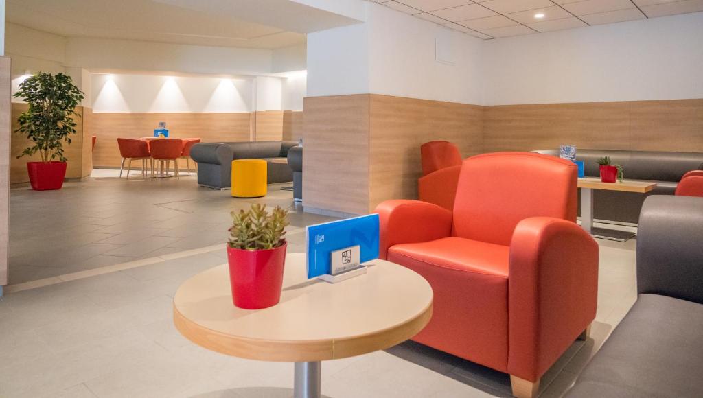 Mūsdienīgs viesnīcas atpūtas zonas interjers ar sarkaniem un pelēkiem krēsliem, koka apdari un dažādām krāsainām mēbelēm. Telpā ir augi un gaismas elementi, kas rada patīkamu atmosfēru.