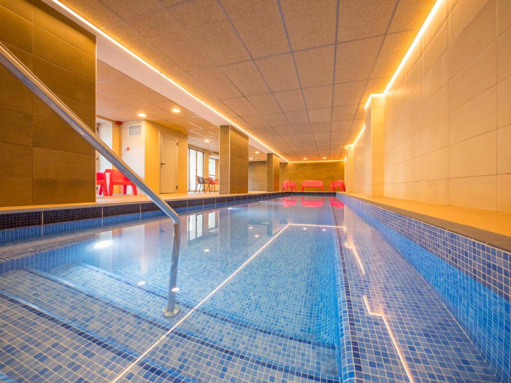 Mūsdienīga iekštelpu peldbaseina telpa ar zilu ūdeni un gaismas efektiem. Interjerā ir sarkanas krēsli un gaišas flīzes, radot relaksējošu atmosfēru.