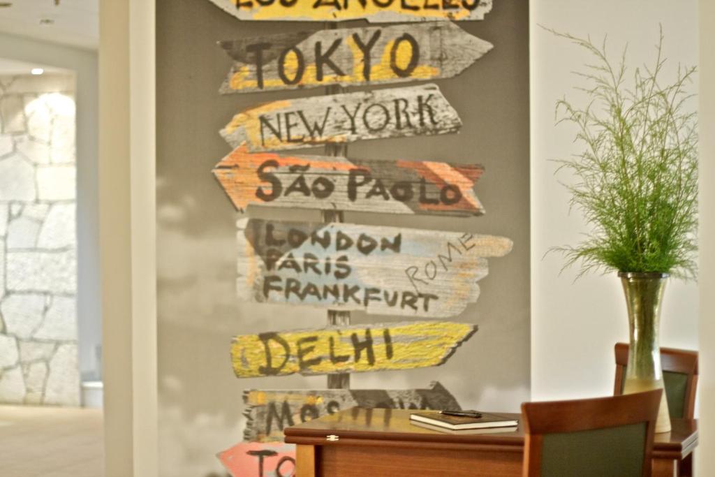 Sienas dekors ar norādēm uz dažādām pilsētām, tostarp Tokiju, Ņujorku un Londonu, radot ceļojumu noskaņu. Priekšplānā redzama koka galds ar ziedu pušķi.