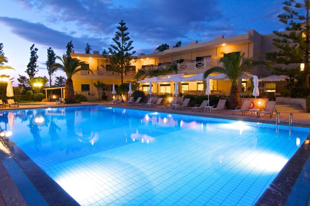 Lielisks nakts skats uz baseinu ar apgaismojumu un viesnīcu, ko ieskauj palmas un zāliens. 