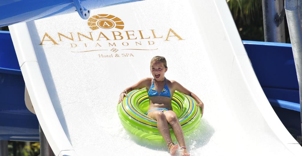Bērns jautri slidinās pa ūdens slidkalniņu ar riņķi, aiz viņa redzama Annabella Diamond viesnīcas un SPA logo.