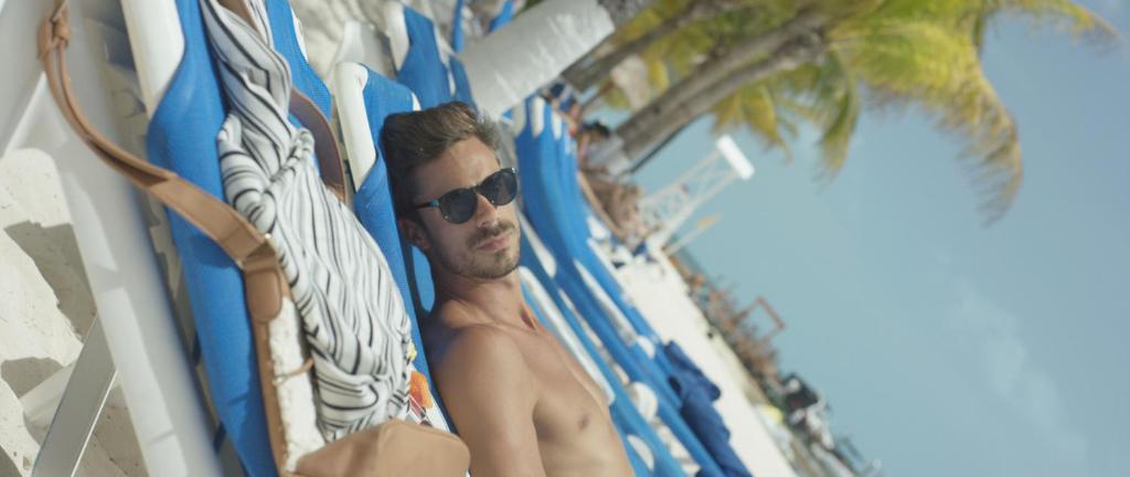 Vīrietis atpūšas pludmalē, guļot uz saulē izkārtām krēslām, ar saulesbrillēm un pludmales fona.