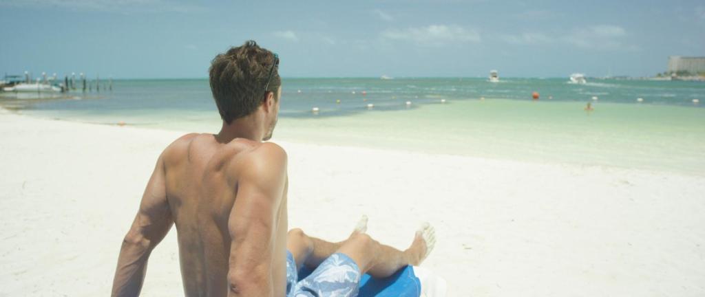 Vīrietis atpūšas pludmalē, skatoties uz jūru, sēžot uz saules krēsla. Apkārt ir balts smiltis un zila ūdens.