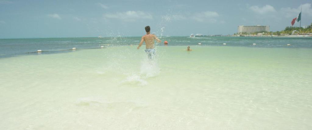 Vīrietis skriežas pa pludmali, izsistot ūdens šļakatas. Fons ietver mierīgu jūru un viesnīcu pie krasta.