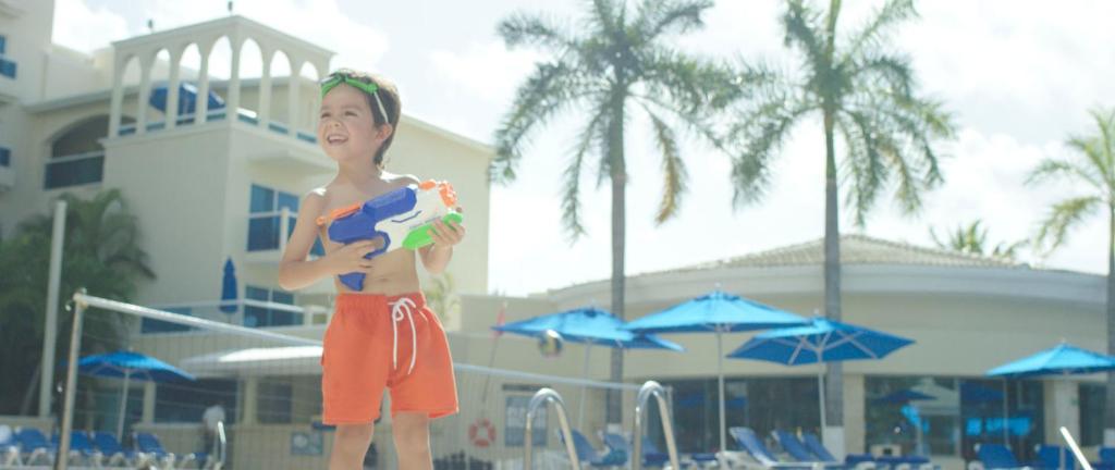 Bērns ar ūdens pistoli, priecīgi stāv pie baseina, apkārt palmām un saules gultām. Ideāla vasaras izklaide pie ūdens.