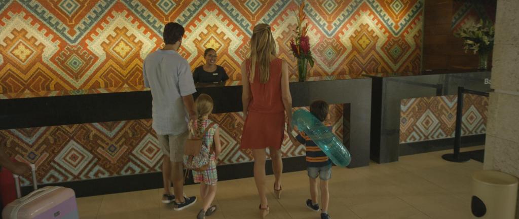 Ģimenes ieeja viesnīcā ar krāsainu mozaīkas fona dizainu un laipnu reģistratūras darbinieku. Bērni tur pludmales piederumus, gatavojoties atpūtai.