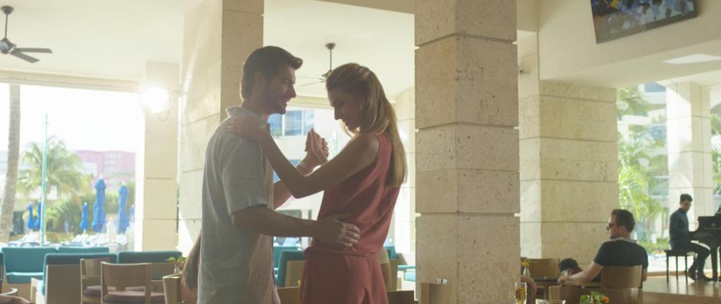 Jaunais pāris dejo viesnīcas atpūtas zonā, apņemti ar gaismu un tropisku atmosfēru. Fonā redzami apmeklētāji un pianists, radot relaksētu noskaņu.