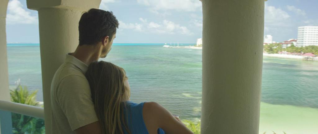 Pāris skatās uz jūru no balkona, baudot mierīgu brīdi kopā. Aiz viņiem redzams skaists piekrastes skats ar zilo ūdeni un palmām.