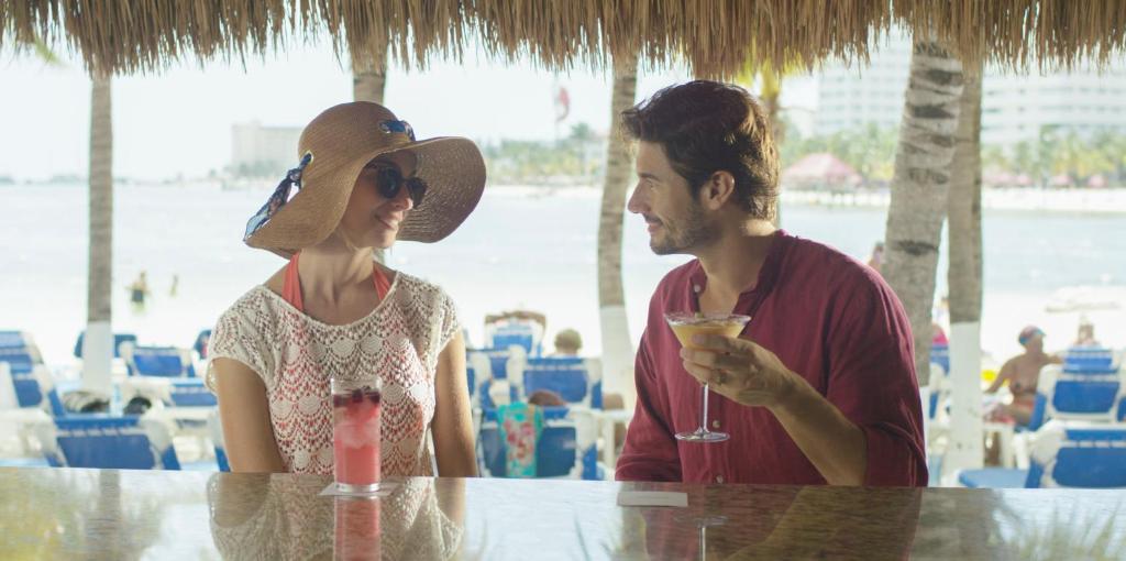Pāris sēž pludmales bārā, baudot kokteiļus un sarunājoties. Fons attēlo pludmali un sauļošanās krēslus, radot relaksējošu atmosfēru.