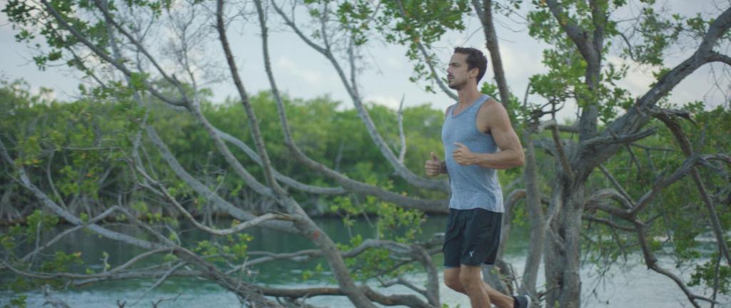 Vīrietis skrien gar krastu, apzaļumotā apkārtnē pie ūdens. Fons ir kluss un dabisks, ar kokiem un zāli.
