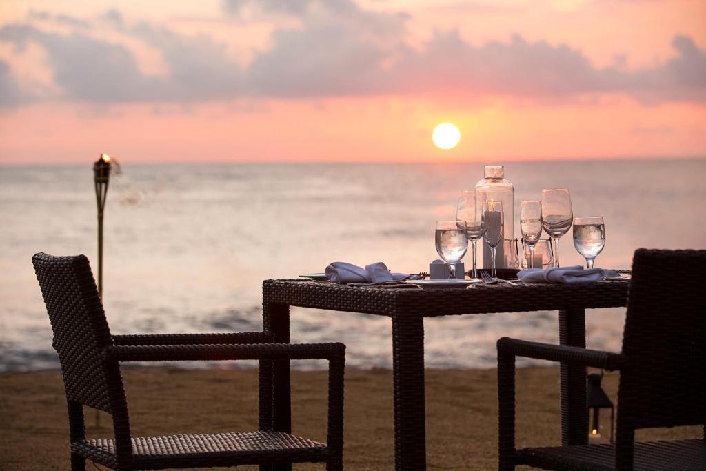 Romantisks vakariņu galds ar skatu uz saulrietu, dekorēts ar stikla traukiem un svečturiem pie jūras. Ideāla atmosfēra romantiskām vakariņām.
