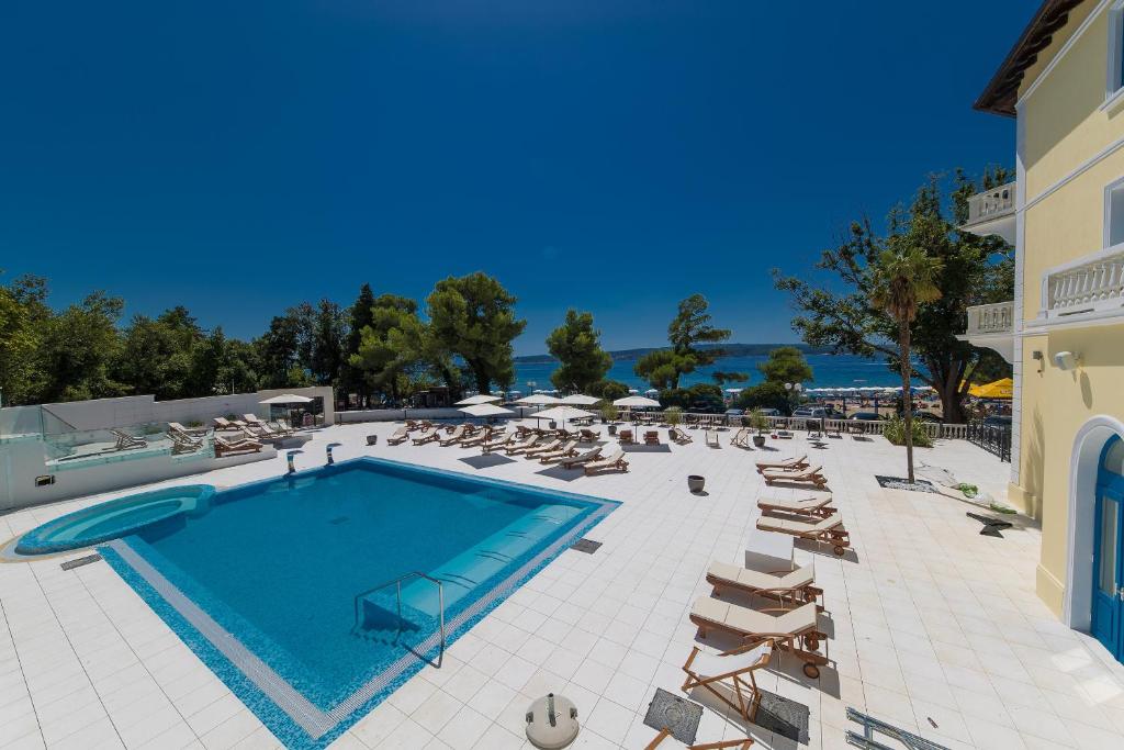 Luksusa viesnīcas baseins ar saulessargiem un atpūtas krēsliem, ieskauts zaļām kokiem un skatu uz jūru. Ideāla vieta atpūtai un relaksācijai.