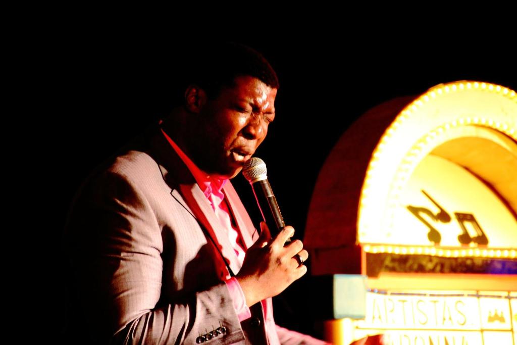 Mākslinieks uzstājas uz skatuves, turot mikrofonu un izrādot emocijas, kamēr fona gaismas izgaismo retro stila mūzikas aparātu.