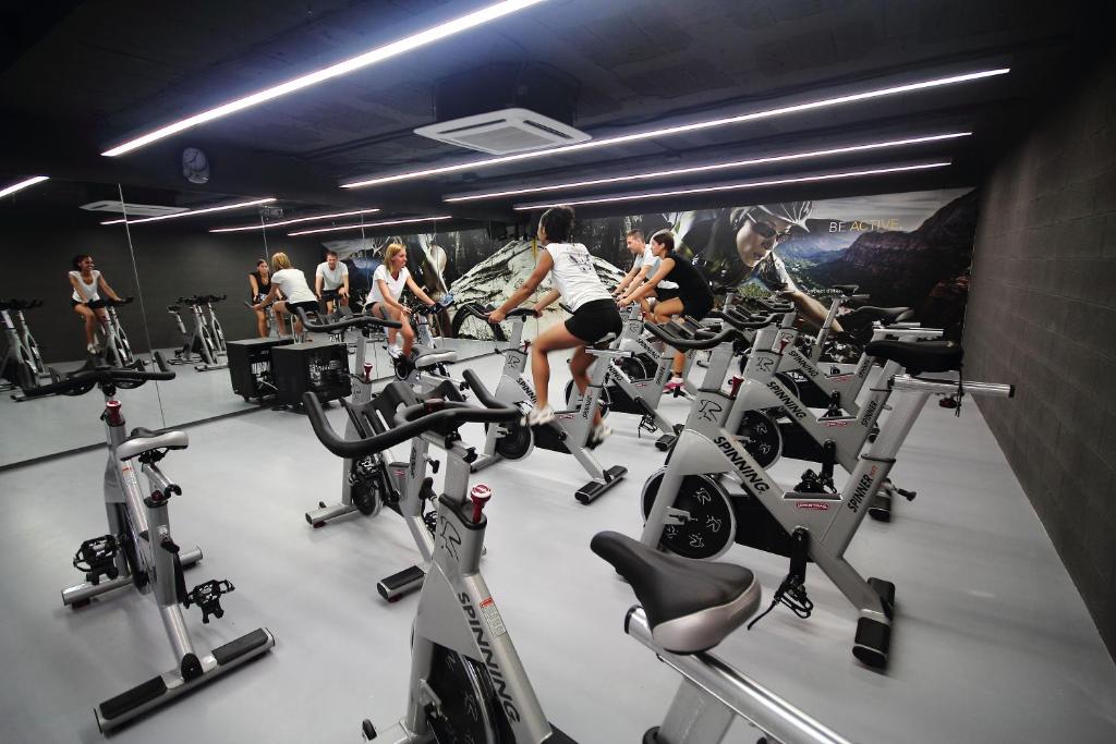 Vingrošanas zāle ar grupas nodarbību, kur dalībnieki trenējas uz stacionārajām velosipēdiem. Moderns interjers ar spoguļiem un iedegtu apgaismojumu.