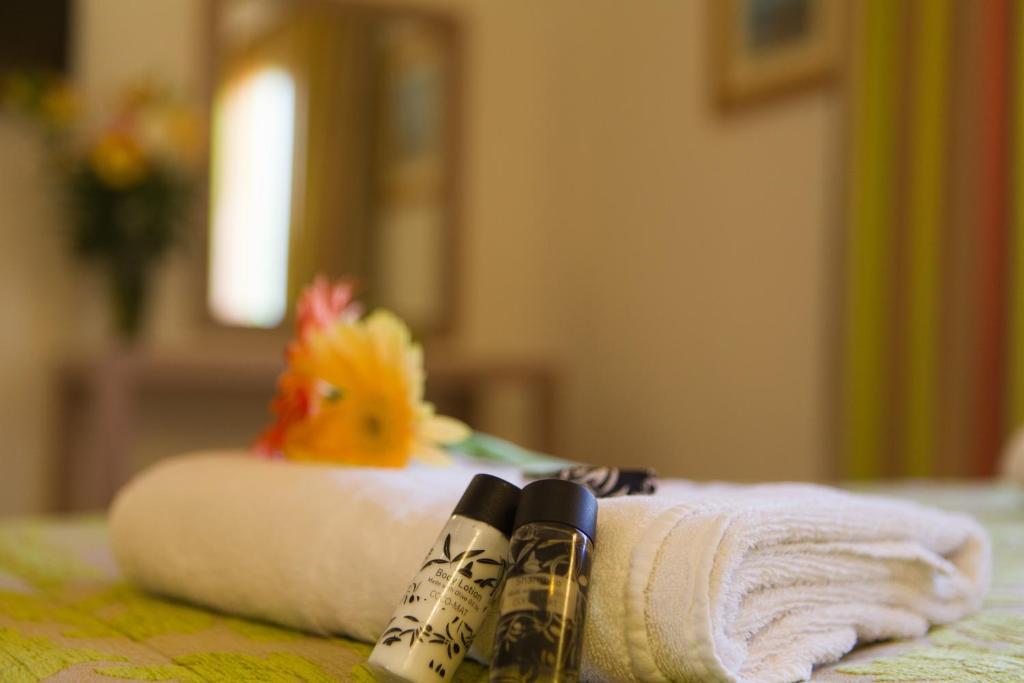 Vannas istabas aksesuāri, tostarp dvielis un divas dažādas ķermeņa kopšanas pudelītes, izvietoti uz gultas. Fons ir mīksts un mājīgs, ar ziediem un krāsainām aizkariem.