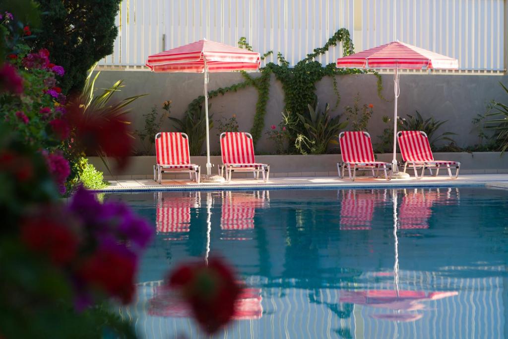 Sarkano un balto striķu sauļošanās krēsli un lietussargi pie peldbaseina, ko ieskauj ziedoši augi un zaļumi.