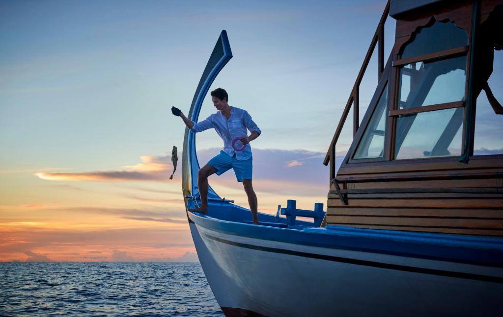 Vīrietis stāv uz laivas, turot roku zivij, fona ir krāšņa saulrieta ainava jūrā.