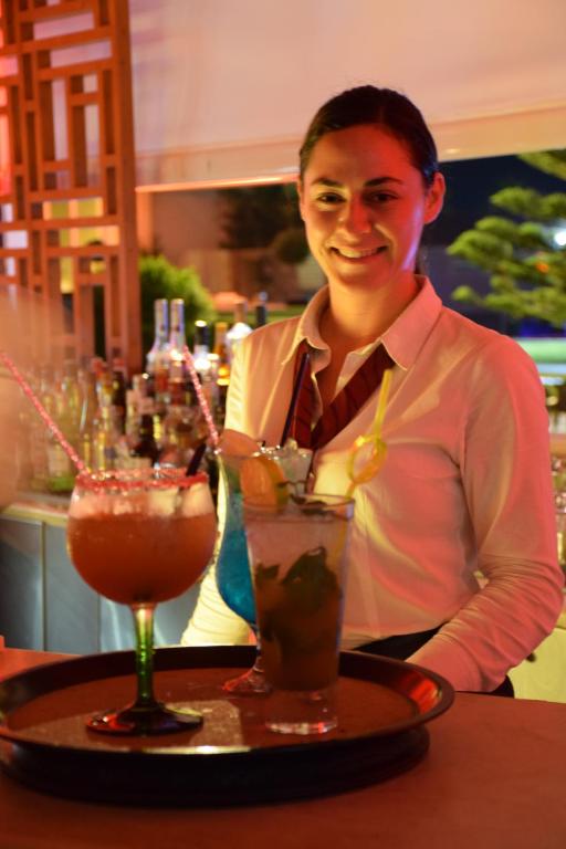 Bārmenis pasniedz divus kokteiļus uz tray, fona apgaismojumā redzama bāra vide. Kokteiļi ir krāsaini un dekorēti ar augļiem un salmiņiem.