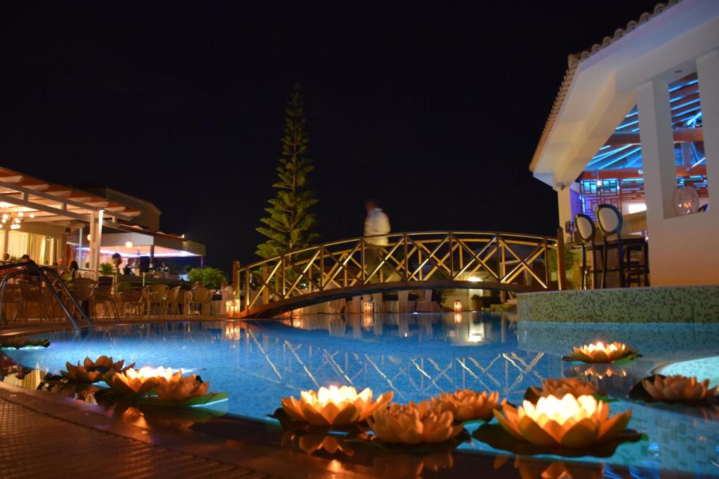 Nakts ainava ar apgaismotu baseinu, ziedošām ūdensrožu lampām un koka tiltu, kas ved uz restorānu. Eleganta atmosfēra ar zāles un apgaismojumu.