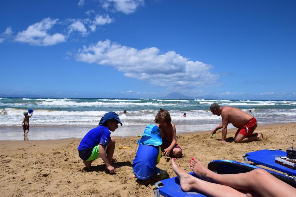 Bērni spēlējas pludmalē, veidojot smilšu pilis, kamēr pieaugušie relaksējas saulē un vēro jūras viļņus. Skaists saulains laiks un zila debesis papildina pludmales atmosfēru.