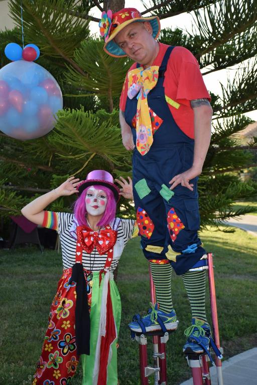 Divi klauni, viens stāv uz stiltēm, otrs ar rozā matiem un košām drēbēm, priecīgā noskaņojumā pasākuma laikā. Fons ar ziediem un gaisa baloniem papildina svētku atmosfēru.