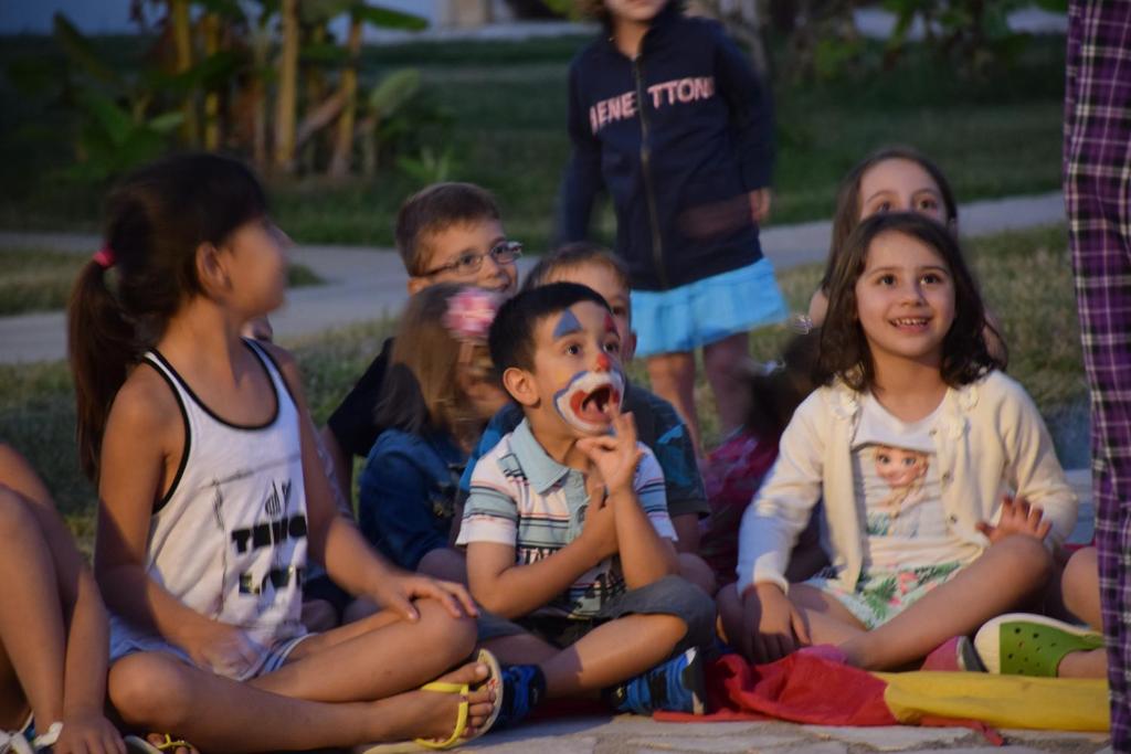 Bērni sēž aplī, skatoties uz izrādi, izrādot prieku un interesi. Daži no viņiem smejas un izskatās apmierināti, radot jautru atmosfēru.