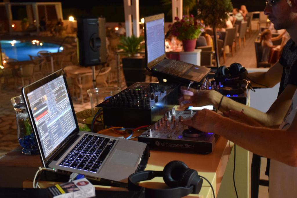 DJ pult ar datoru un audio aprīkojumu, kas izmantots nakts pasākumā pie baseina, radot atmosfēru izklaidei. Fons ietver apgaismotu baseinu un viesus, kas bauda vakaru.