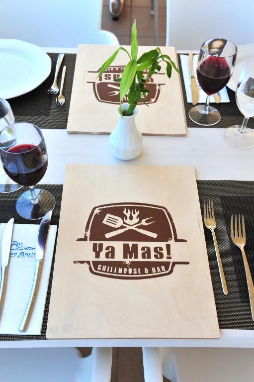 Galda klājums ar koka ēdienkarti "Ya Mas! Grill House & Bar", kas papildināts ar sarkano vīnu un dekoratīvu augu.
