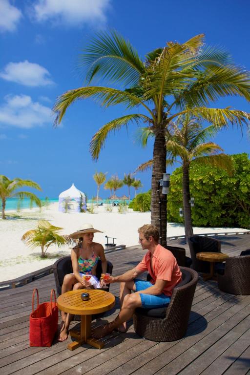 Pāris atpūšas tropu pludmalē, sēžot ērtos krēslos pie koka galda, baudot dzērienus un saules gaismu. Fons ietver palmām un zilo jūras ainavu.