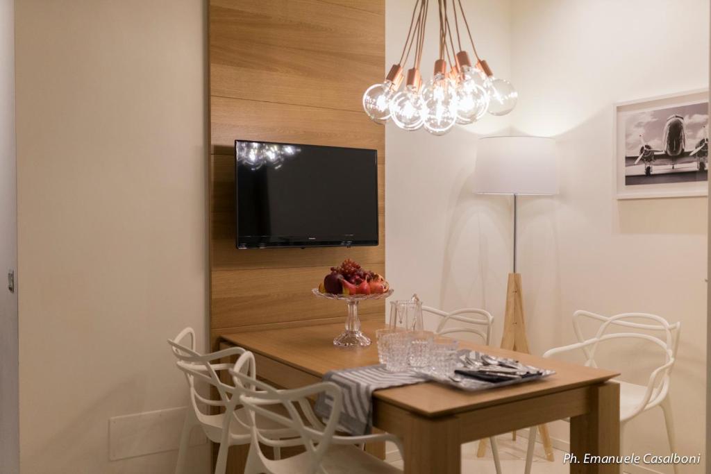 Mūsdienīgs ēdamistabas interjers ar koka galdu, baltām krēsliem un modernu gaismekli. Uz galda atrodas augļu kompozīcija un galda piederumi.