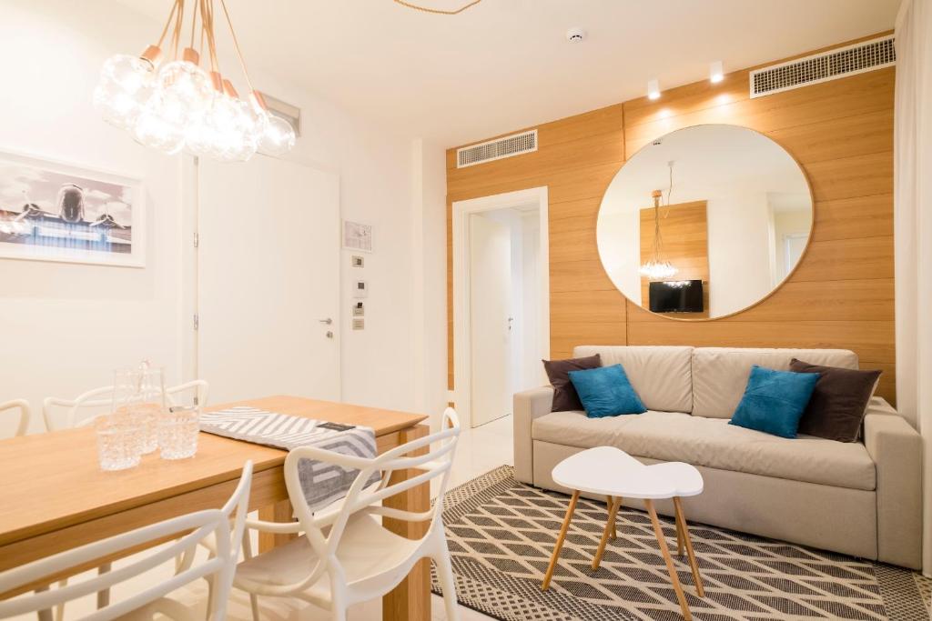 Mūsdienīgs viesistabas un ēdamistabas interjers ar gaišiem toņiem, koka mēbelēm un stilīgu apgaismojumu. Uz dīvāna ir spilveni, bet uz galda ir glāzes un galdauts.