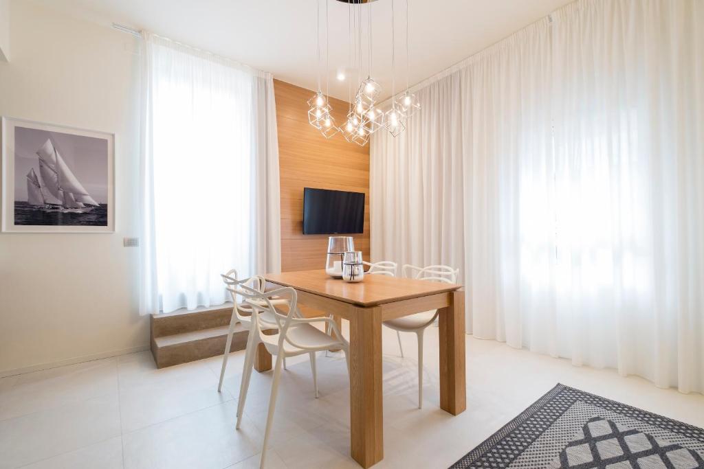 Moderns ēdamistabas interjers ar koka galdu un baltām krēsliem, apgaismots ar stilīgu lampu. Logi ar vieglām aizkarām nodrošina dabīgu apgaismojumu.