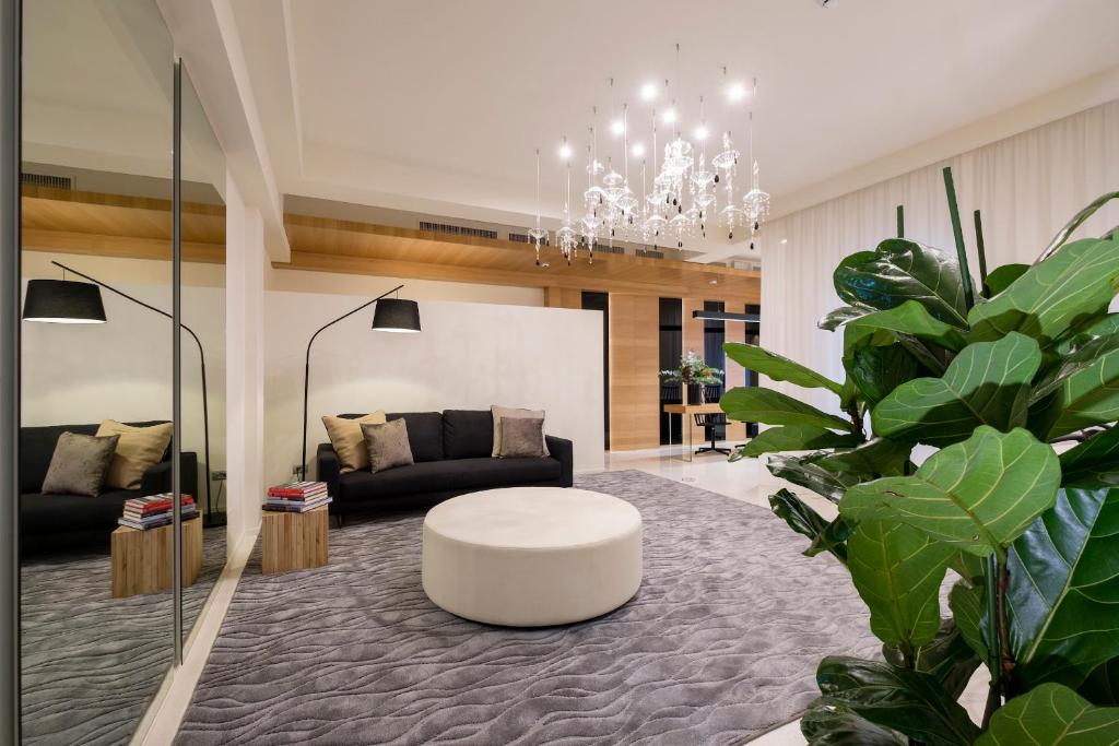 Mūsdienīgs dzīvojamās istabas interjers ar melnu dīvānu, apaļu kafijas galdiņu un elegantu lustras apgaismojumu. Telpā ir liels spogulis un dekoratīvi augi, radot mājīgu atmosfēru.