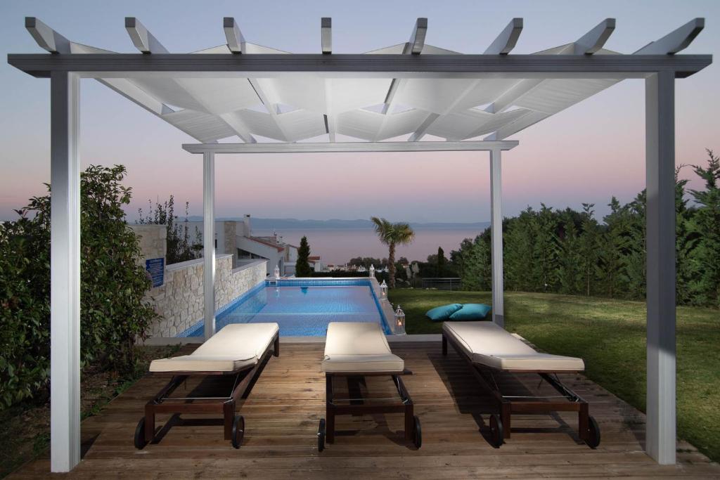 Mūsdienīgs atpūtas stūrītis ar divām saulessargu gultām zem balta nojumes, blakus peldbaseinam un zaļiem krūmiem, ar skatu uz jūru un saulrietu.