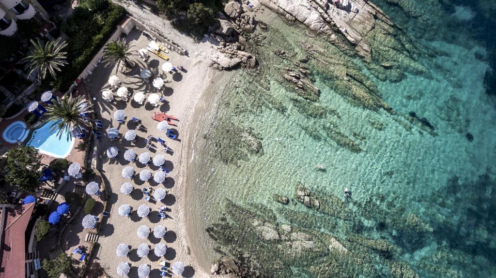 Pludmales skats no augšas ar saulessargiem, peldētājiem un skaidru ūdeni. Apkārt palmām un baseinam, kas veido relaksējošu atpūtas vidi.