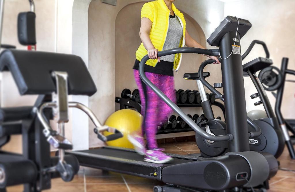 Sieviete vingro sporta zālē uz eliptiskā trenažiera, ar dzeltenu vestīti un rozā legingiem, fona redzami svaru stieņi un citi trenažieri.