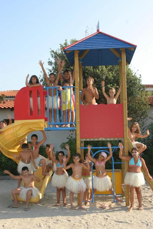 Bērni priecīgi spēlējas uz rotaļu laukuma, tērpušies apģērbos no salmiem, un izsaka prieku, stāvot pie slidkalniņa. Fons ir saulains, un apkārt ir zaļumi.