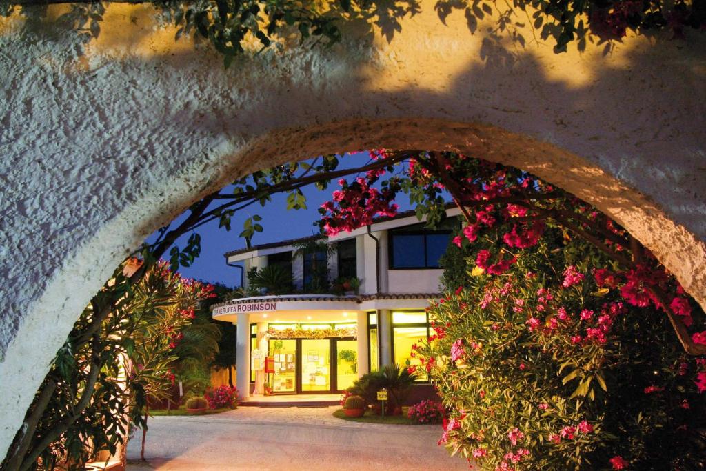 Skaists viesnīcas ieejas skats, apvīts ar ziedošām krūmiem un dārzu, kas izgaismots maigos nakts toņos.