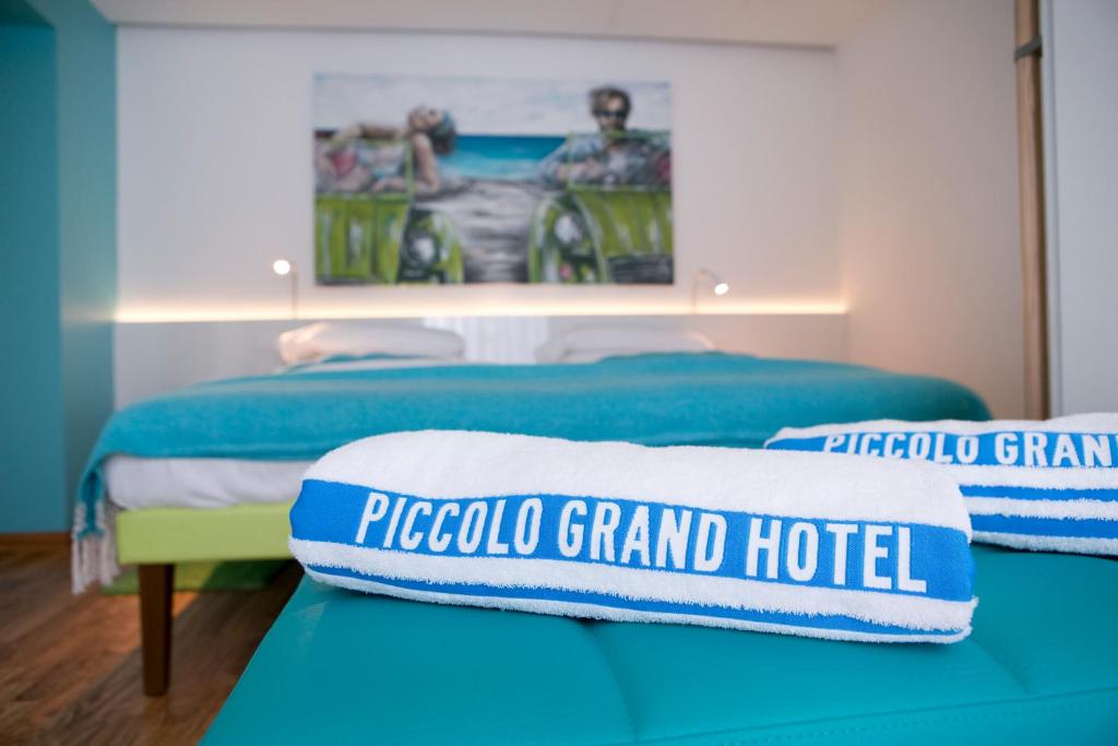 Mūsdienīga viesnīcas numura interjers ar gulošām gultām un spilgtiem dvieļiem, uz kuriem redzams “PICCOLO GRAND HOTEL” nosaukums. Fons rotā gleznainu jūras ainu.