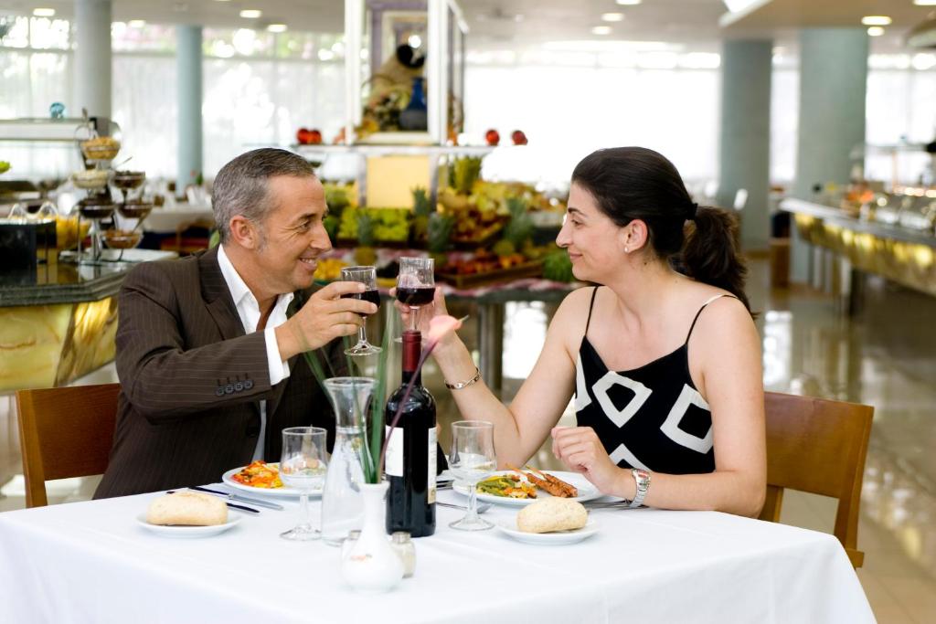 Romantiska vakariņas diviem, kur pāris smaidot, pacelt glāzes sarkanvīna, baudot gardu ēdienu elegantā restorānā. Gaisīga atmosfēra ar sulīgiem augļiem fonā.