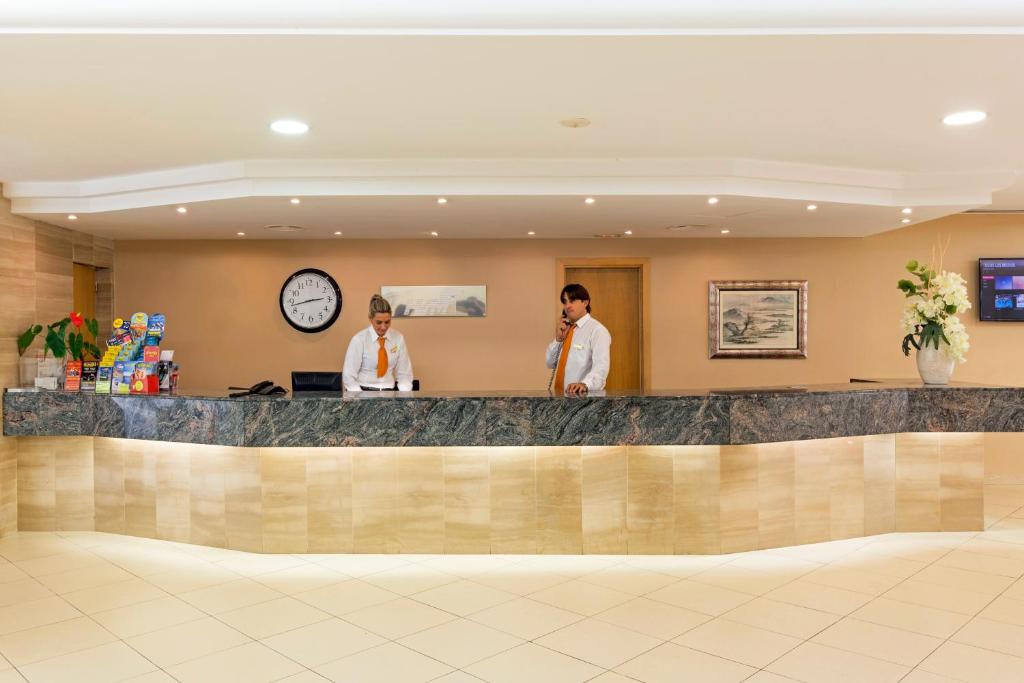 Viesnīcas reģistratūra ar diviem darbiniekiem, no kuriem viens runā pa telefonu, bet otrs strādā pie datora. Uz galda atrodas brošūras un ziedi.