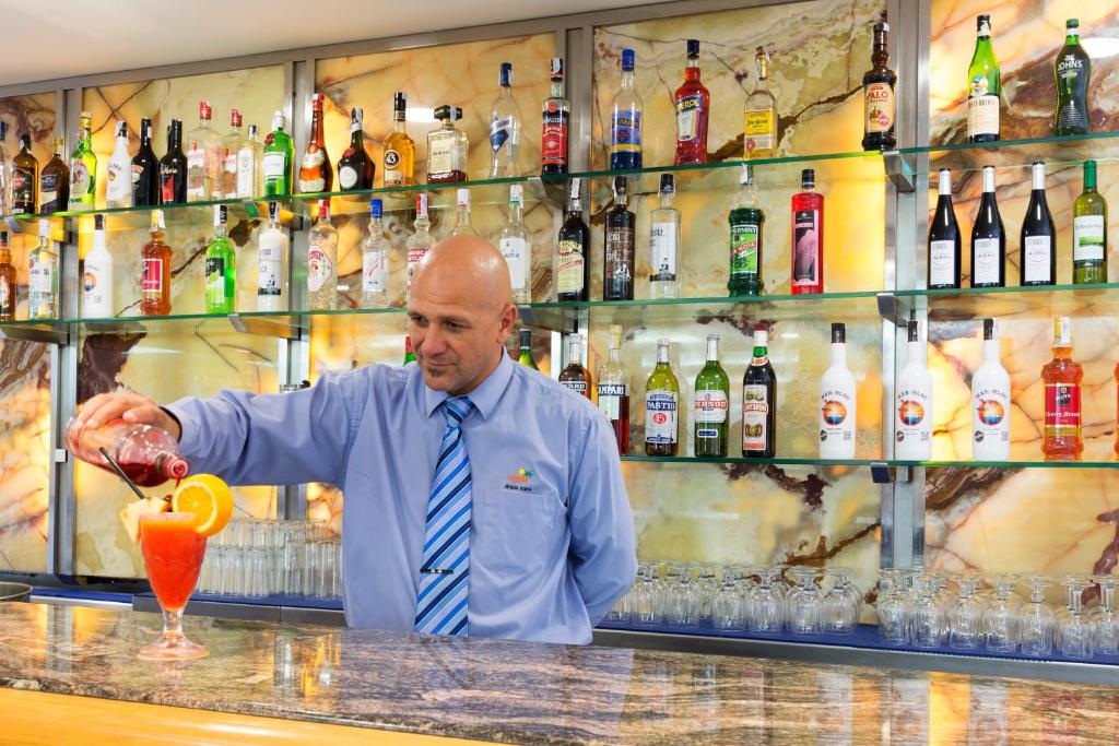 Bārmenis pie bāra ielej kokteili ar apelsīnu un sarkano sīrupu, fona plauktā redzami dažādi alkoholiskie dzērieni.